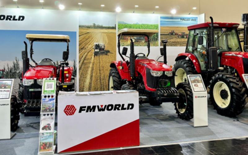 Stan pameran pertanian menampilkan tiga traktor—ukuran kompak, sedang, dan besar—di tengah pemandangan lahan pertanian, dengan branding 'FMWORLD' yang menonjol.