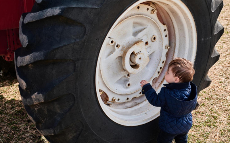 Anak kecil berbaju biru di samping ban traktor besar, tampak penasaran